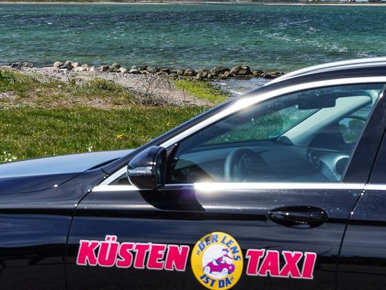 Küsten-Taxi mit Schriftzug auf der Fahrertür, im Hintergrund die Ostsee